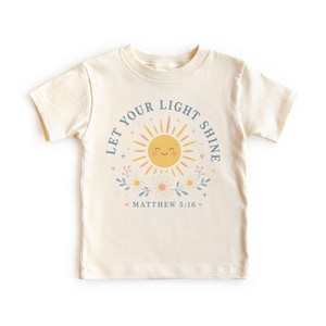 May include: A cream-coloured short-sleeve t-shirt featuring a graphic of a smiling yellow sun surrounded by flowers and the text "LET YOUR LIGHT SHINE" in blue, along with the scripture reference "MATTHEW 5:16" printed in a matching blue font.