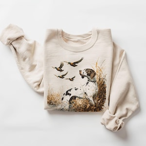 Puede incluir: Una sudadera beige con un estampado gráfico de un perro marrón y blanco con patos volando por encima. El perro está sentado en hierba alta.