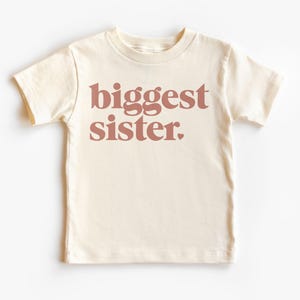 May include: A cream colored t-shirt with short sleeves and the words "biggest sister" printed in a pink color on the front.