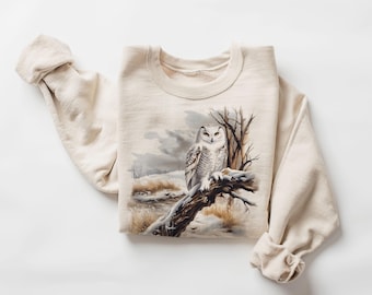 Könnte beinhalten: Ein beiger Pullover mit einer Schnee-Eule, die auf einem Ast in einer winterlichen Szene sitzt. Die Eule hat weiße Federn mit schwarzen Markierungen und gelbe Augen.