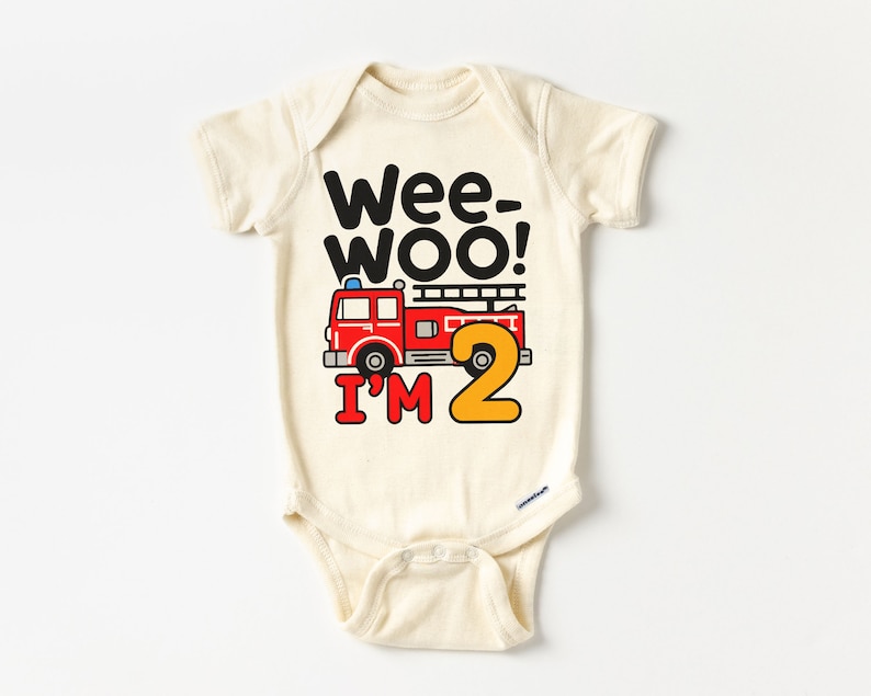 May include: A cream-colored baby onesie with a red fire truck and the text "Wee-Woo! I'm 2" in black and yellow.
