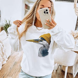 May include: A white long-sleeve crewneck sweatshirt featuring a graphic print of a grey heron bird wearing a mustard yellow knit beanie. The bird is depicted in profile facing left with detailed feathers and a long, pointed yellow beak.