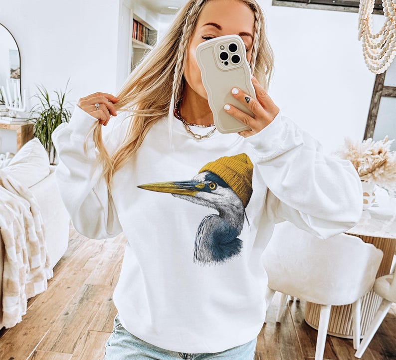 May include: A white long-sleeve crewneck sweatshirt featuring a graphic print of a grey heron bird wearing a mustard yellow knit beanie. The bird is depicted in profile facing left with detailed feathers and a long, pointed yellow beak.