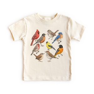 以下が含まれることがあります： さまざまな鳥のカラフルなイラストが描かれた、クリーム色のTシャツ。赤いカージナル、ルリビタキ、黄色いフィンチなどが含まれています。デザインはシャツの前面に配置されています。