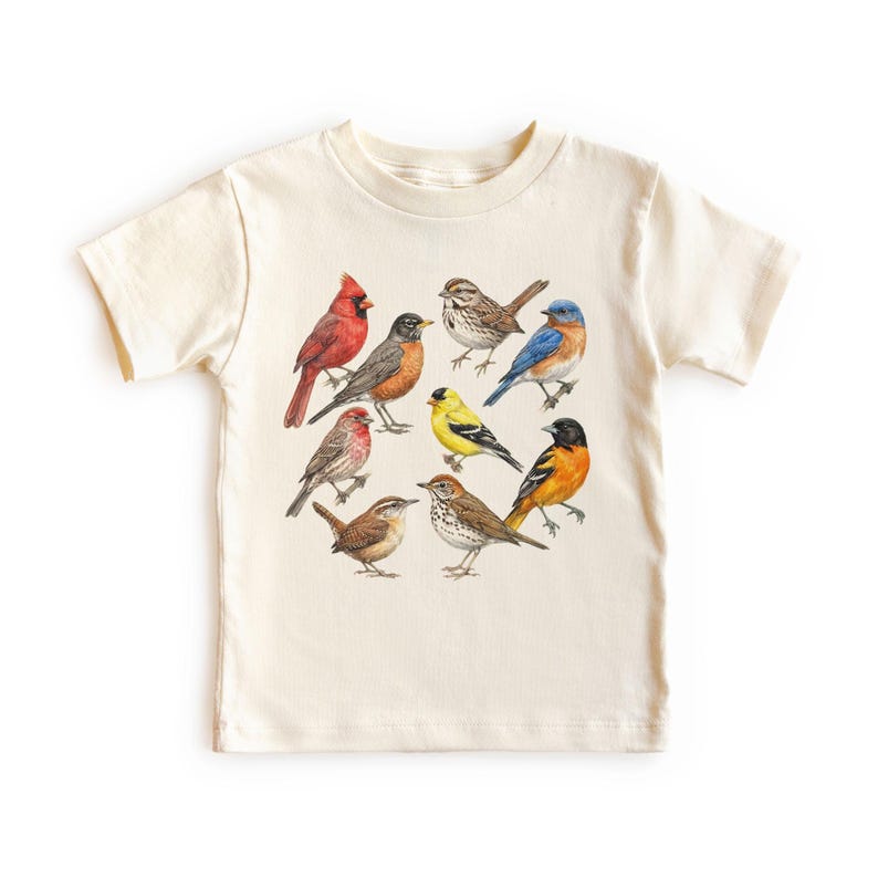 以下が含まれることがあります： さまざまな鳥のカラフルなイラストが描かれた、クリーム色のTシャツ。赤いカージナル、ルリビタキ、黄色いフィンチなどが含まれています。デザインはシャツの前面に配置されています。