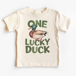 Puede incluir: Camiseta color crema con una ilustraci&oacute;n de acuarela de un pato real. El texto "ONE LUCKY DUCK" est&aacute; impreso en verde encima y debajo del pato. La camiseta est&aacute; hecha de un material suave.