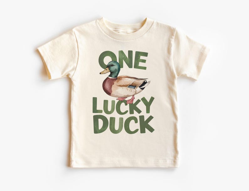 Puede incluir: Camiseta color crema con una ilustraci&oacute;n de acuarela de un pato real. El texto "ONE LUCKY DUCK" est&aacute; impreso en verde encima y debajo del pato. La camiseta est&aacute; hecha de un material suave.