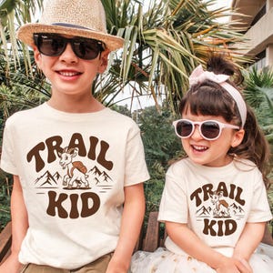 Peut inclure: Deux enfants portant des t-shirts cr&egrave;me avec le texte "TRAIL KID" en marron, ainsi qu'un dessin de ch&egrave;vre et de montagnes. L'un porte un chapeau de paille et des lunettes de soleil, l'autre porte des lunettes de soleil et un bandeau rose.