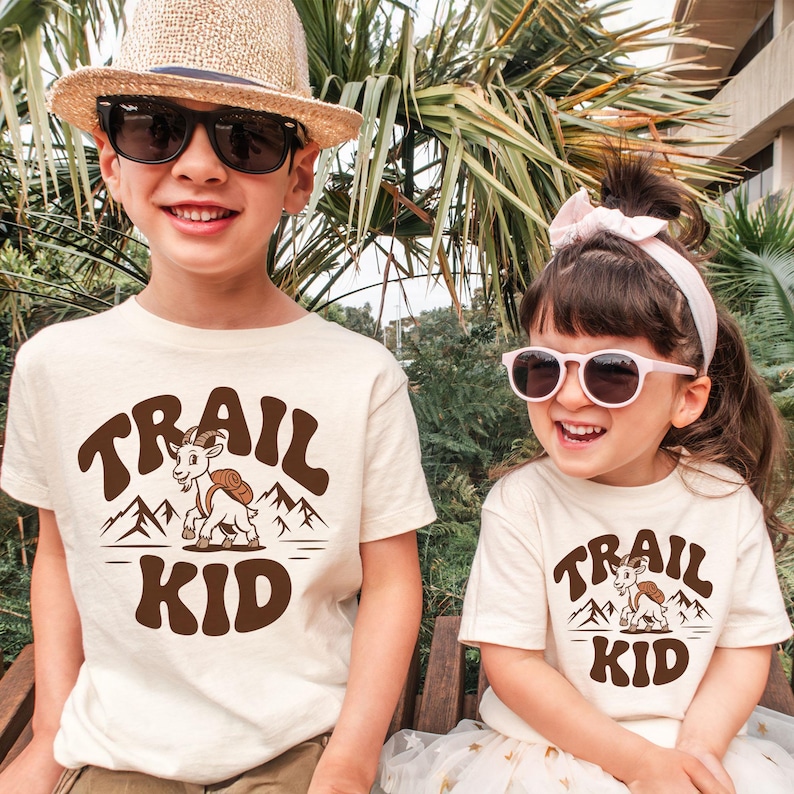 Peut inclure: Deux enfants portant des t-shirts cr&egrave;me avec le texte "TRAIL KID" en marron, ainsi qu'un dessin de ch&egrave;vre et de montagnes. L'un porte un chapeau de paille et des lunettes de soleil, l'autre porte des lunettes de soleil et un bandeau rose.