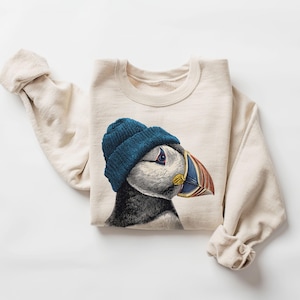 May include: A cream-coloured sweatshirt with a detailed illustration of a puffin wearing a blue beanie. The puffin has a colourful beak with red, blue, and yellow accents. The sleeves are rolled up, and the design is casual and comfortable.