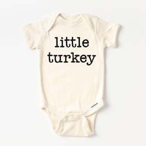 May include: A cream-colored baby bodysuit with the words "little turkey" printed in bold black letters. The short-sleeved onesie has a round neckline and snap closures at the bottom.