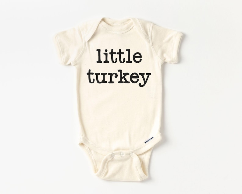 May include: A cream-colored baby bodysuit with the words "little turkey" printed in bold black letters. The short-sleeved onesie has a round neckline and snap closures at the bottom.