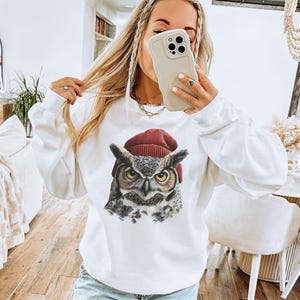 May include: A white long-sleeve sweatshirt featuring a graphic print of a great horned owl wearing a dark red knit beanie. The owl has detailed brown, gray, and white feathers with piercing yellow eyes, centered on the chest of the garment.