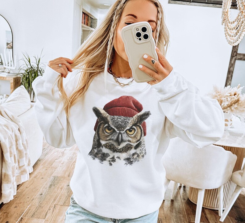 May include: A white long-sleeve sweatshirt featuring a graphic print of a great horned owl wearing a dark red knit beanie. The owl has detailed brown, gray, and white feathers with piercing yellow eyes, centered on the chest of the garment.