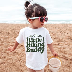 May include: A white t-shirt with the text "Little Hiking Buddy" in dark green, featuring mountain and tree graphics. The child is wearing pink sunglasses and holding a yellow bucket on a sandy beach.