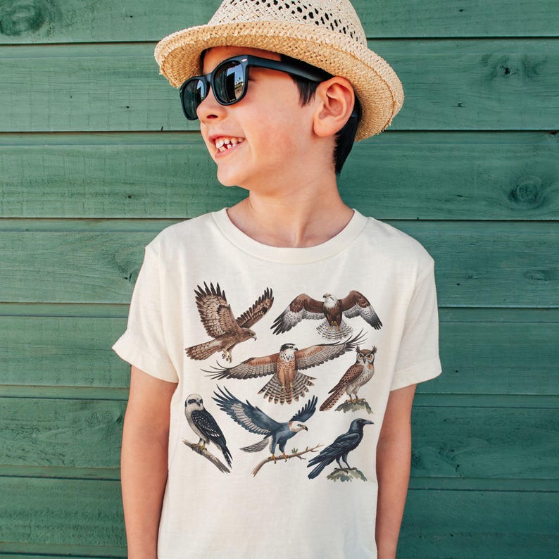 K&ouml;nnte beinhalten: Ein cremefarbenes T-Shirt mit Illustrationen verschiedener Greifv&ouml;gel, darunter Falken, Adler und Eulen. Das T-Shirt wird von einer Person getragen, die einen Strohhut und eine Sonnenbrille tr&auml;gt.