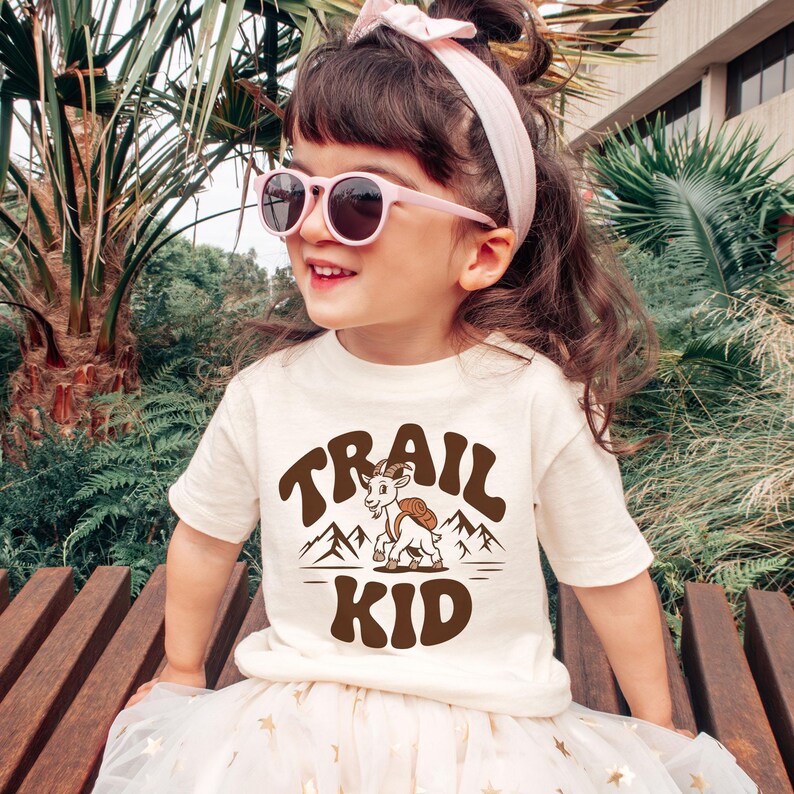 Peut inclure: Un enfant portant un t-shirt cr&egrave;me avec les mots "TRAIL KID" en marron, ainsi qu'un dessin de ch&egrave;vre et de montagnes. L'enfant porte &eacute;galement des lunettes de soleil roses et un bandeau rose. L'enfant est assis sur un banc en bois.