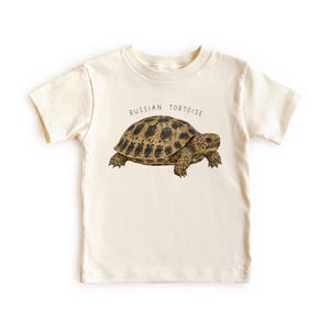 May include: A cream-colored short-sleeve t-shirt featuring a detailed illustration of a Russian tortoise in shades of brown and black. The text "RUSSIAN TORTOISE" is printed in a simple, dark font in an arc above the tortoise illustration.