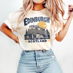 Pu&ograve; includere: Una t-shirt a maniche corte color crema con una stampa grafica blu scuro dello skyline di Edimburgo, in Scozia, che include il castello e un grande sole giallo. Il testo riporta Edinburgh sopra la citt&agrave; e Scotland sotto con delle linee.