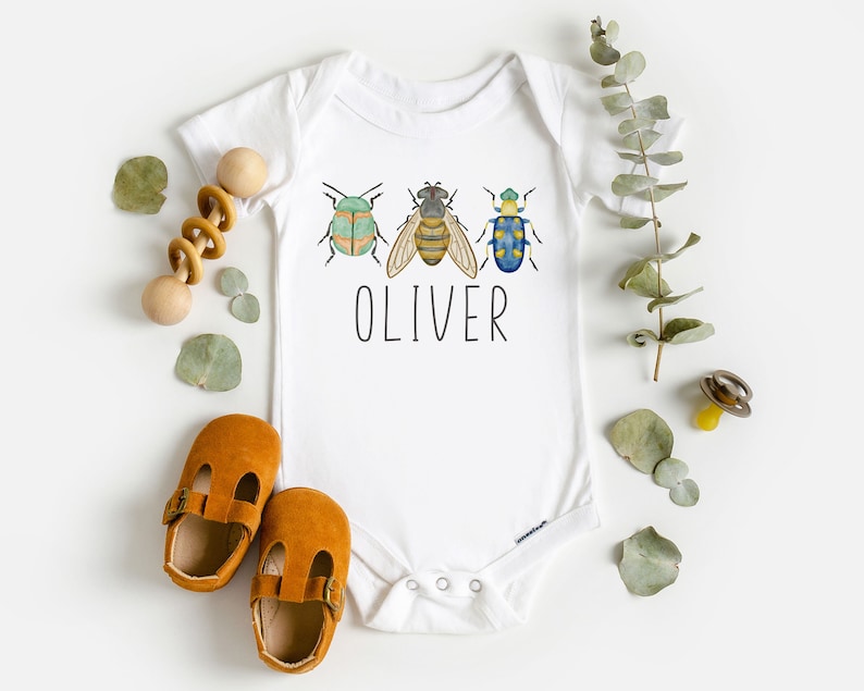 May include: White baby onesie with a green, yellow, and blue bug design and the name "OLIVER" printed in black.