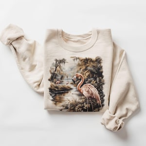 May include: A beige sweatshirt featuring a printed design of pink flamingos in a swampy landscape.