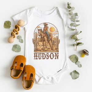 May include: White baby onesie with a brown cowboy riding a horse in a desert scene with a yellow sun and cacti. The text "HUDSON" is printed in brown below the image.