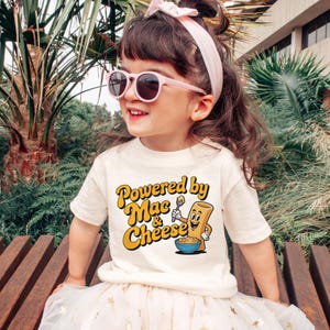 K&ouml;nnte beinhalten: Ein Kind tr&auml;gt ein cremefarbenes T-Shirt mit dem Text "Powered by Mac & Cheese" in Gelb und Orange. Das Shirt zeigt eine Cartoon-Figur von Mac and Cheese. Das Kind tr&auml;gt auch eine rosa Sonnenbrille und ein rosa Stirnband.