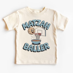 Peut inclure: T-shirt crème avec un motif sur le thème du basket-ball. Le motif comprend les mots "MATZAH BALLER" en lettres bleues, une boulette de matzah de dessin animé, un panier de basket et un bol. Un design amusant et ludique pour enfants.