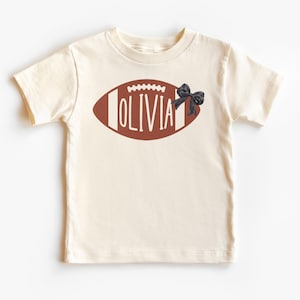 Puede incluir: Camiseta color crema con un dise&ntilde;o de bal&oacute;n de f&uacute;tbol americano marr&oacute;n y el nombre "OLIVIA" en letras blancas. Un lazo negro est&aacute; en el lado derecho del bal&oacute;n. La camiseta es de manga corta.