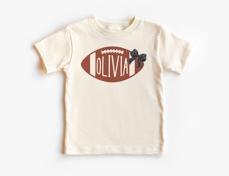Puede incluir: Camiseta color crema con un dise&ntilde;o de bal&oacute;n de f&uacute;tbol americano marr&oacute;n y el nombre "OLIVIA" en letras blancas. Un lazo negro est&aacute; en el lado derecho del bal&oacute;n. La camiseta es de manga corta.