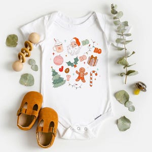 Puede incluir: Body de beb&eacute; blanco con un dise&ntilde;o navide&ntilde;o colorido. El dise&ntilde;o incluye a Pap&aacute; Noel, un &aacute;rbol de Navidad, un hombre de jengibre y la frase "HO HO". El body est&aacute; rodeado de zapatos de beb&eacute; y un juguete de madera.