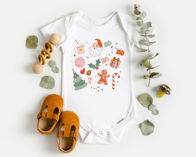 Puede incluir: Body de beb&eacute; blanco con un dise&ntilde;o navide&ntilde;o colorido. El dise&ntilde;o incluye a Pap&aacute; Noel, un &aacute;rbol de Navidad, un hombre de jengibre y la frase "HO HO". El body est&aacute; rodeado de zapatos de beb&eacute; y un juguete de madera.