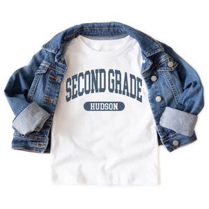 May include: A white t-shirt with the text "SECOND GRADE HUDSON" in navy blue, with a blue denim jacket draped over it. The jacket has silver buttons and a classic design.
