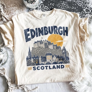 Pu&ograve; includere: Una t-shirt a maniche corte color crema con una stampa grafica blu scuro dello skyline di Edimburgo in Scozia. Il design include un grande sole giallo dietro il castello, con il testo Edinburgh sopra e Scotland sotto l'illustrazione.