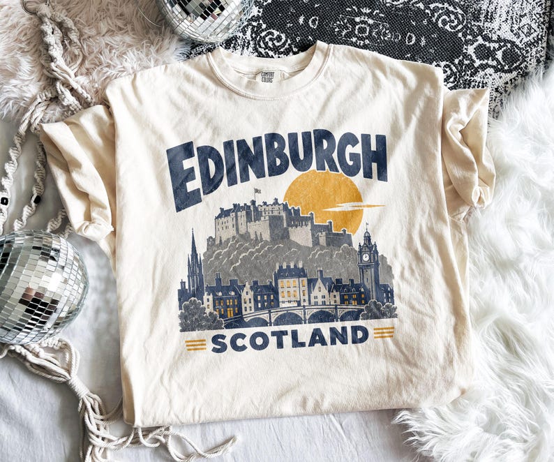 Pu&ograve; includere: Una t-shirt a maniche corte color crema con una stampa grafica blu scuro dello skyline di Edimburgo in Scozia. Il design include un grande sole giallo dietro il castello, con il testo Edinburgh sopra e Scotland sotto l'illustrazione.