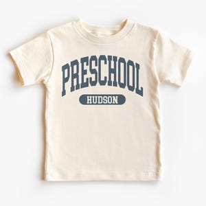 May include: Cream-colored short-sleeved t-shirt with the word "PRESCHOOL" in navy blue arched across the chest. Below the word "PRESCHOOL" is the word "HUDSON" in navy blue on a navy blue rectangular background.