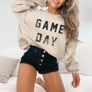 May include: A beige sweatshirt with the words "GAME DAY" printed in black. The sweatshirt is paired with black denim shorts. The image is taken indoors.