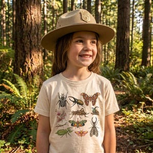 Pode incluir: Uma pessoa jovem usando um chapéu de guarda florestal bege com um distintivo e uma camiseta bege com ilustrações coloridas de insetos. O fundo é uma exuberante floresta verde com árvores altas.