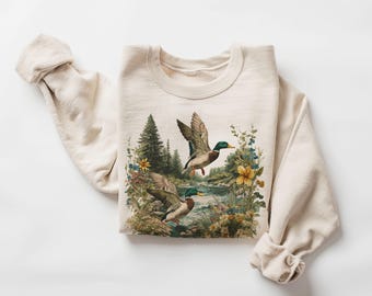 May include: Cream-colored sweatshirt featuring a nature scene with two ducks in flight over a river, surrounded by flowers and trees. The long sleeves are cuffed at the wrists. The graphic has a vintage, painterly style.
