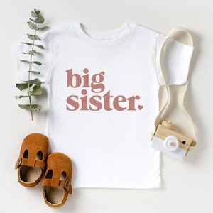May include: White t-shirt with the words "big sister" printed in pink on the front.