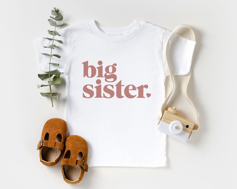 May include: White t-shirt with the words "big sister" printed in pink on the front.