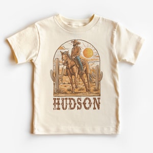 May include: A beige t-shirt with a graphic of a cowboy riding a horse in a desert landscape with cacti and a sun. The text "HUDSON" is printed below the graphic.