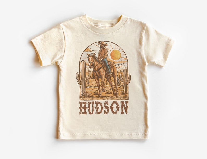 May include: A beige t-shirt with a graphic of a cowboy riding a horse in a desert landscape with cacti and a sun. The text "HUDSON" is printed below the graphic.