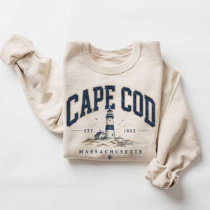 Op de afbeelding: Een crèmekleurige sweatshirt met de marineblauwe tekst "CAPE COD" in een boog. Daaronder een vuurtorenillustratie met de tekst "MASSACHUSETTS" en "EST. 1602". De mouwen zijn opgerold.
