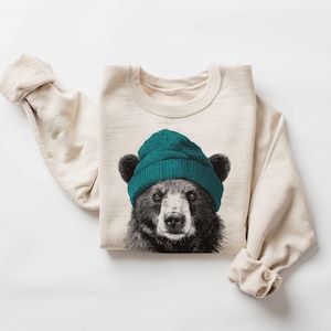 Op de afbeelding: Crèmekleurige sweatshirt met een zwart-wit berenillustratie met een tealkleurige beanie. De lange mouwen zijn opgerold en het gezicht van de beer staat in het midden van de voorkant. De sweatshirt is gemaakt van zacht materiaal.