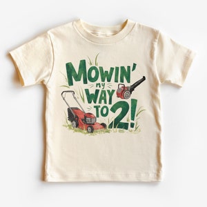 May include: A light beige t-shirt with a green and red illustration of a lawnmower and leaf blower. The text "Mowin' my way to 2!" is printed on the shirt.