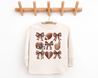 May include: A cream-coloured long-sleeved shirt adorned with watercolour illustrations of football-themed elements. The design includes brown bows, a football, a helmet, a foam finger, and a heart-shaped football.