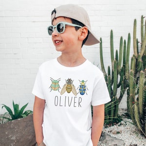 May include: A white t-shirt with a green, yellow, and blue bug design and the name "Oliver" printed on the front. The shirt is being worn by a child wearing a light blue baseball cap and sunglasses.