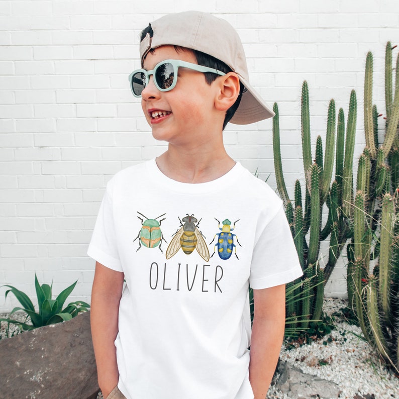 May include: A white t-shirt with a green, yellow, and blue bug design and the name "Oliver" printed on the front. The shirt is being worn by a child wearing a light blue baseball cap and sunglasses.