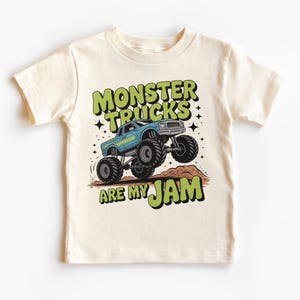 Puede incluir: Camiseta color crema con un gr&aacute;fico de cami&oacute;n monstruo azul y el texto "MONSTER TRUCKS ARE MY JAM" en verde y negro. El dise&ntilde;o incluye estrellas y un mont&iacute;culo de tierra marr&oacute;n.